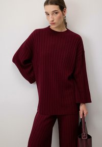 Bordový pletený svetr s širokými rukávy, spárovaný s odpovídajícími kalhotami. Modelka drží ladící kabelku.