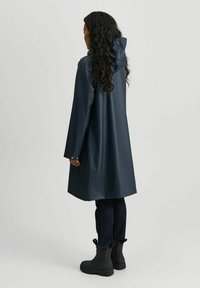 Marineblå vanntett regnfrakk med hette, lange ermer og trykknapper på mansjettene, sammen med svarte boots og mørke jeans. Glatt, glansfull tekstur.