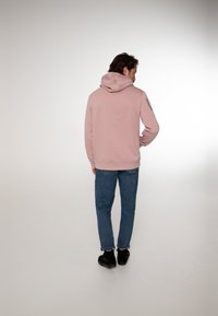 Felpa rosa con tasca a marsupio frontale, polsini e orlo a coste. Indossata con jeans in denim blu e scarpe nere, vista da dietro.