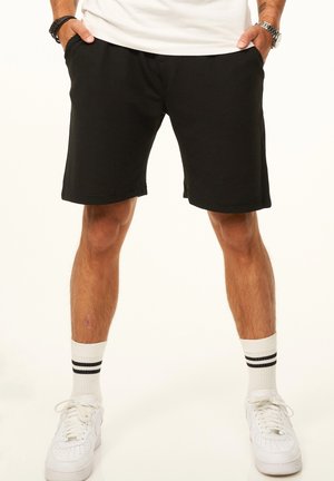 Mann trägt schwarze Shorts, weiße Sportsocken mit schwarzen Streifen und weiße Sneakers, steht mit den Händen in den Taschen vor einem einfarbigen Hintergrund.