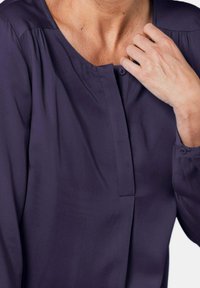 Blusa blu navy con scollatura rotonda, chiusura a bottoni e maniche lunghe. Presenta un tessuto morbido e pieghe sottili vicino alle spalle.