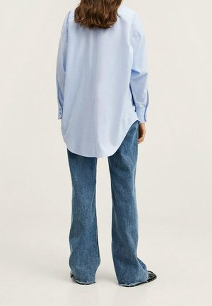 Person med langt brunt hår iført en lyseblå oversized skjorte og vidbenet blå jeans, stående foran en ensfarvet baggrund.