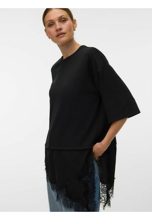 Femme portant un haut noir ample à manches courtes avec un ourlet en dentelle sur un jean bleu clair, debout avec une main dans la poche sur fond blanc.