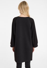 Schwarze Tunika mit einer gesteppte Textur auf dem Rücken, Fledermausärmeln und lockerer Passform. Glattes Material mit matter Oberfläche, das bis zur Mitte des Oberschenkels reicht.