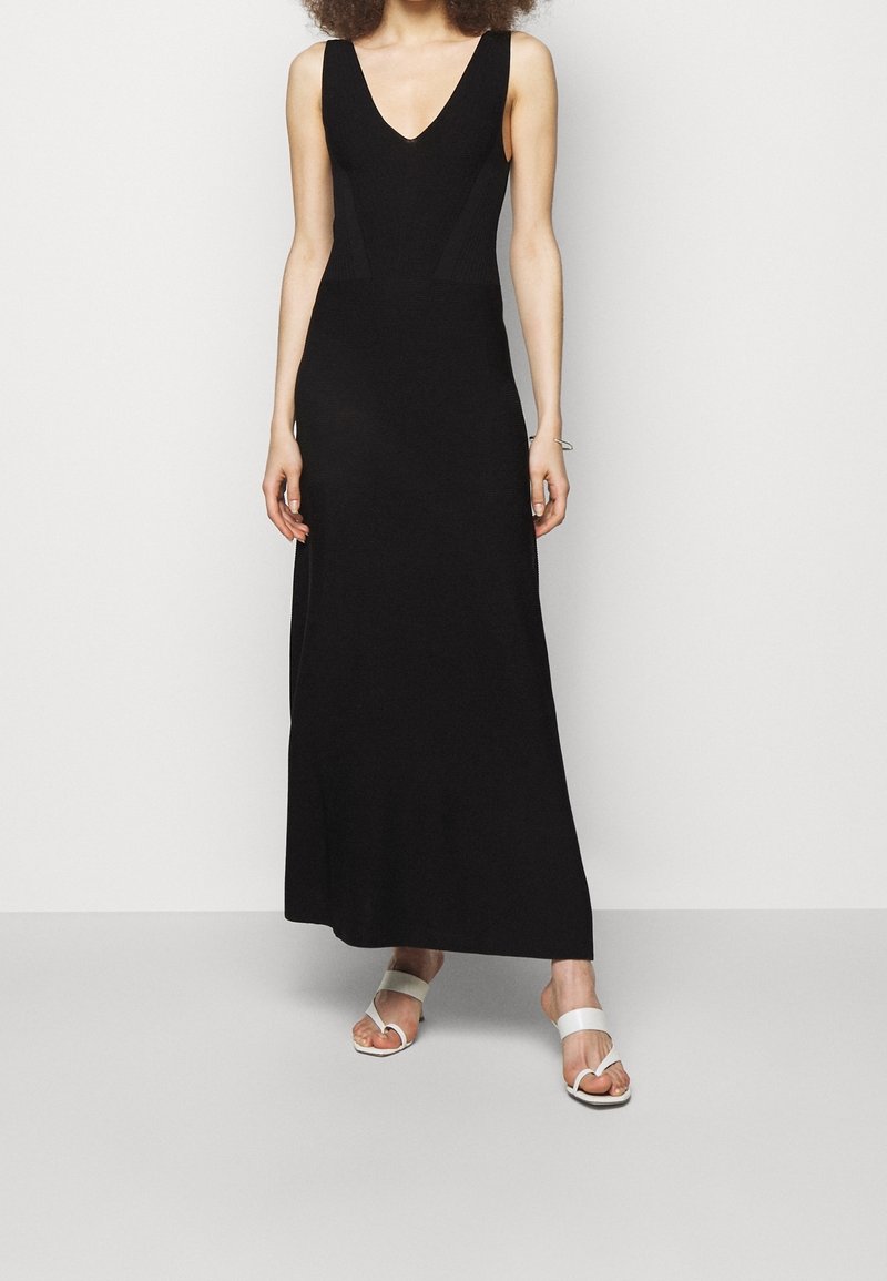 Svart stickad maxiklänning med djup V-ringning och sidofickor. Bärs med vita sandaler med breda remmar och en slät, platt sula.