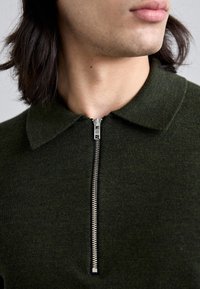 Polo de punto verde oscuro con una cremallera corta en el cuello y un cuello clásico, con un acabado de tela texturizada.