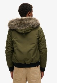 Persona che indossa un giubbotto con cappuccio verde oliva con bordi in pelliccia sintetica e pantaloni beige, di spalle contro uno sfondo bianco.