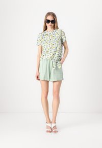 T-shirt branca com padrão floral e mangas curtas, atada na cintura; combinada com calções verdes claros; modelo usa óculos de sol pretos e sandálias brancas.