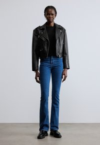 Geacă de piele neagră pentru motocicletă, cu fermoare argintii, asortată cu blugi evazați albaștri și pantofi negri, pe un model pe fundal neutru.