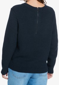 Vrouw in een marineblauwe ribgebreide trui met een ritssluiting op de rug en lichtblauwe jeans, gezien van achteren.