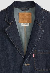 Μαύρο τζην μπλέιζερ με καπιτονάτο πέτο, τσέπη στο στήθος, κίτρινες ραφές και δερμάτινη ετικέτα με λεπτομέρειες της μάρκας μέσα στον γιακά.