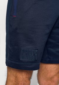 Shorts de sport bleu marine en tissu lisse, avec un patch logo texturé et des surpiqûres contrastantes près de l'ourlet.