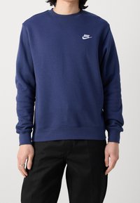 Person som bär en marinblå Nike-tröja med logotyp på vänster bröst och svarta byxor, ståendes mot en enkel ljus bakgrund.