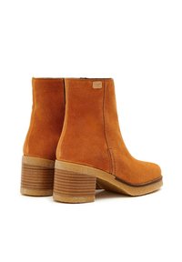 Botas de ante color óxido hasta el tobillo, con un tacón de madera apilado, punta redonda y detalles de costura sutiles. Presenta una pequeña etiqueta de la marca en el costado.