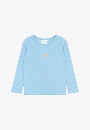 Abito a salopette blu navy con orlo arricciato, abbinato a una maglietta a maniche lunghe azzurra e scarpe verdi, accentuato da calzini con motivo floreale.