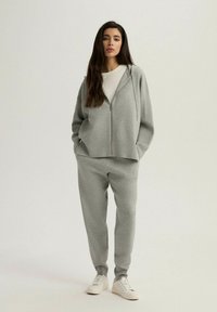Szara bluza z kapturem na zamek z kieszeniami, w zestawie z dopasowanymi dresami. Miękka tkanina, luźny krój, biała koszulka pod spodem i białe tenisówki.