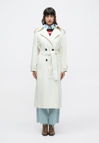 Trenchcoat i lysecremefarvet stof med bælte, dobbeltradede sorte knapper, skulderklaffer og knælængde design. Blå denimjeans og brune støvler.