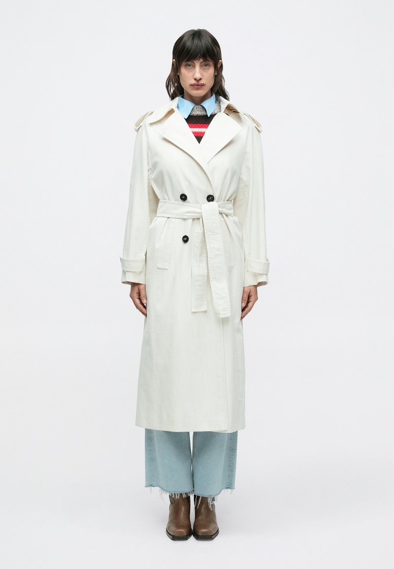 Trenchcoat i lysecremefarvet stof med bælte, dobbeltradede sorte knapper, skulderklaffer og knælængde design. Blå denimjeans og brune støvler.