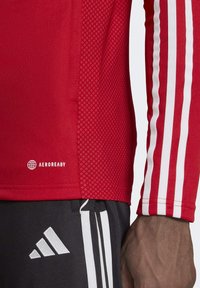 Rotes sportliches Langarmshirt mit strukturierten Ärmeln, weißen Streifen und einem AEROREADY-Logo nahe dem Saum. Getragen mit schwarzen Shorts.