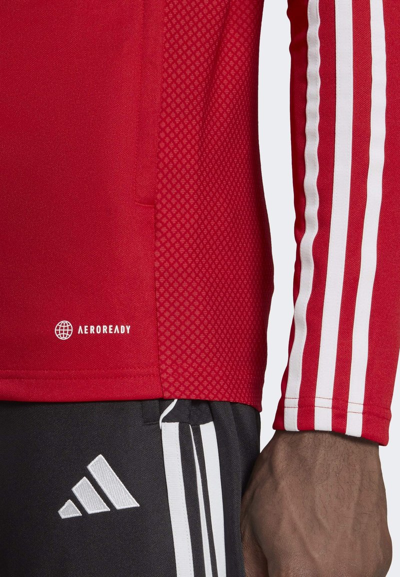 Camiseta atlética roja de manga larga con mangas texturizadas, franjas blancas y un logo AEROREADY cerca del dobladillo. Combinada con pantalones cortos negros.