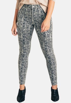 Kobieta w wysokich, dopasowanych spodniach z szaro-czarnym wzorem w skórę węża, zestawionych z czarnymi botkami i beżową bluzką.