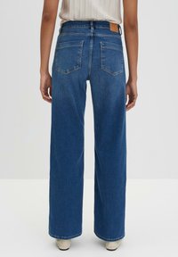 Hoge taille, wijde jeans in donkerblauwe denim. Beschikt over twee achterzakken en een bruine leren patch op de taille. Rechte, schone zoom.