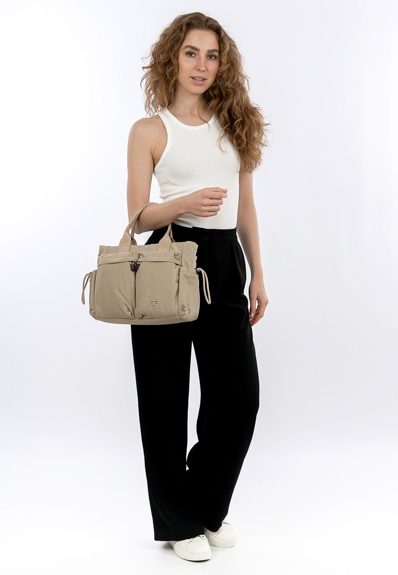 Frau mit lockigem Haar, die ein weißes Tanktop, schwarze Hose mit weitem Bein und weiße Schuhe trägt, hält eine beige Tragetasche mit Vorderfächern und Griffen.