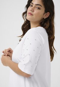 T-shirt blanc à manches courtes avec des clous en argent embellis sur les manches. Tissu en coton lisse, coupe décontractée, col rond et texture douce.