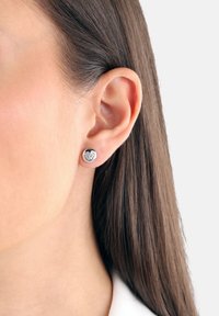Pendiente de studs en forma de corazón de plata con un diseño texturizado, sobre un fondo claro, llevado en la oreja izquierda.