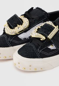 Μαύρα sneakers με υφή, λευκές σόλες, χρυσά καρφιά στα δάχτυλα και μεγάλες χρυσές αγκράφες διακοσμημένες με διαυγείς στρας στα λουριά.