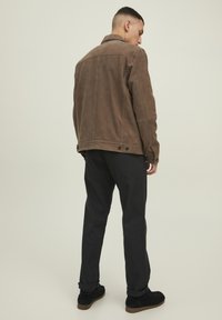 Chaqueta de ante marrón con cuello y puños abotonados, combinada con pantalones negros y zapatos oscuros. La chaqueta tiene un acabado texturizado.