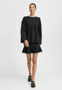 Schwarzes, locker sitzendes Langarm-Oberteil mit Rundhalsausschnitt, kombiniert mit einem schwarzen, schwingenden Miniröckchen. Das Outfit zeichnet sich durch einen strukturierten Stoff und subtile Nähte aus.