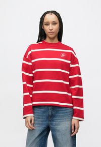 Sweat-shirt à manches longues rayé rouge et blanc avec un col rond et un logo brodé. Porté avec un jean bleu taille haute.