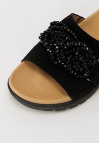 Sandale negre din suede, cu o curea texturată decorată cu mărgele negre. Talpa este bej, cu o ușoară curbă pentru confort.