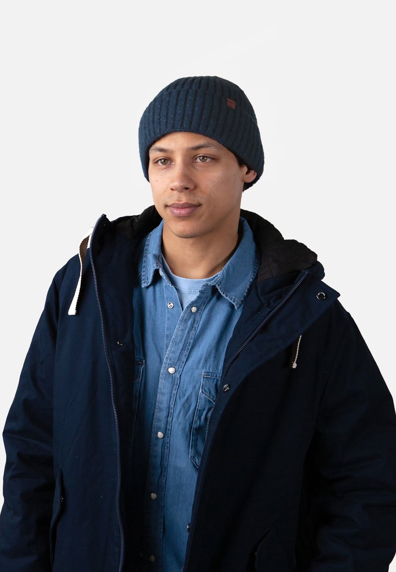 Beanie in maglione navy con texture a coste, abbinato a una giacca scura e a una camicia in denim azzurro chiaro, che presenta dettagli con bottoni e uno stile a strati.
