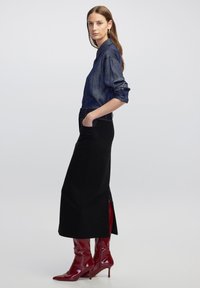 Denim overhemd in donkerblauw, lange mouwen, gecombineerd met een zwarte rok met hoge zijsplitten. Glanzende rode kniehoge laarzen maken de look compleet.