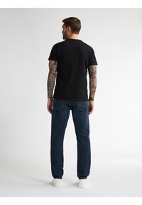 Camiseta negra de manga corta, vista trasera, combinada con jeans azul oscuro. Diseño sencillo, corte regular y zapatillas blancas completan el atuendo.