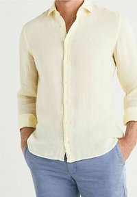 Camisa de lino amarillo claro con mangas largas, frente abotonado y puños enrollados. Presenta una textura sutil y un pequeño detalle bordado.
