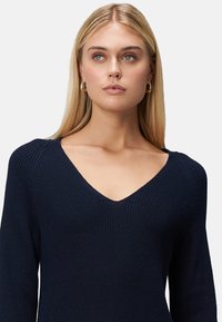 Femme blonde aux cheveux longs portant des boucles d'oreilles en or et un pull en maille bleu marine à col en V, regardant légèrement vers la droite sur un fond uni.
