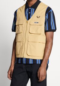 Ljusbeige väst i slitstarkt material, med flera framfickor, en central dragkedja och en svart logotypapplikation. Bärs över en randig skjorta.