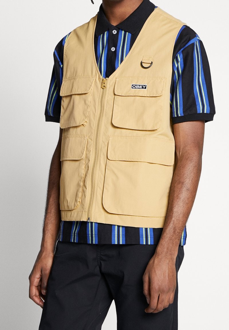 Ljusbeige väst i slitstarkt material, med flera framfickor, en central dragkedja och en svart logotypapplikation. Bärs över en randig skjorta.