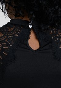 Blouse noire avec des détails en dentelle sur les épaules, un dos ajouré avec deux boutons décoratifs, et un tissu texturé.