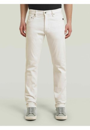 Mann trägt cremefarbene Jeans und graue Sneakers und steht vor einem schlichten hellen Hintergrund.