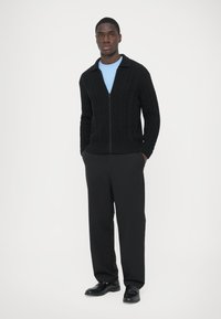 Cardigan noir en maille avec motifs torsadés, fermeture éclair et col, porté sur une chemise bleu clair, associé à un pantalon noir et des chaussures noires.