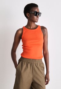 Top deține un decolteu rotund, de culoare portocalie, cu o textură ribbed, asociat cu pantaloni bej cu talie elastică. Ochelarii de soare au un cadru negru cu detalii ale logo-ului.