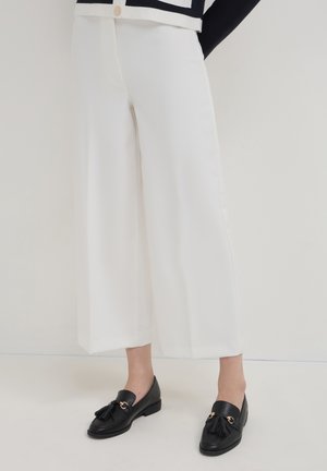White wide-leg pants made of smooth fabric, paired with black loafers featuring gold hardware and a tassel detail.