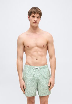 Jaunas vyras su trumpais ruda plaukais, dėvintis šviesiai žalius maudymosi šortus su raštu, stovintis priešais vienspalvį šviesų foną.