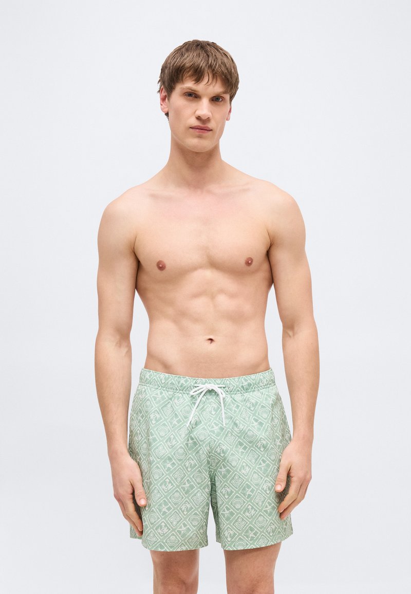 Νεαρός άνδρας με κοντά καστανά μαλλιά, φοράει ανοιχτό πράσινο μαγιό με σχέδια, στέκεται μπροστά σε απλό ανοιχτό φόντο.