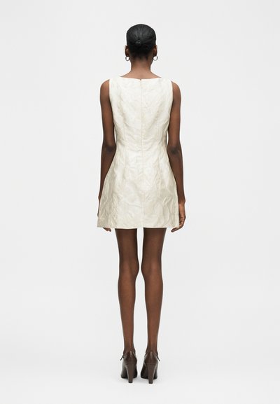 Γυναίκα που στέκεται γυρισμένη πλάτη, φοράει λευκό μίνι φόρεμα χωρίς μανίκια με υφή και καφέ παπούτσια με τακούνι, τα μαλλιά της πιασμένα σφιχτό κότσο.