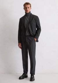 Charcoalgrijze pak met een op maat gemaakte blazer met notch-lapels en bijpassende broek. Draag een grijze coltrui en zwarte geklede schoenen.